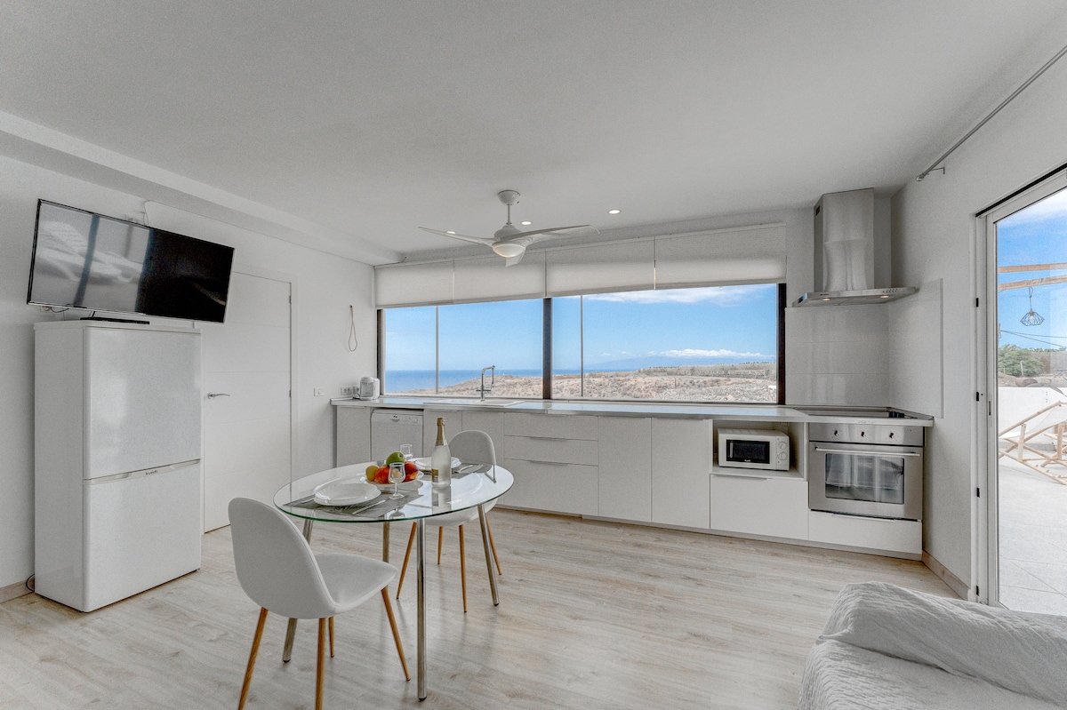 A modern kitchen and dining area are displayed, featuring a circular glass table set with fresh fruit. White cabinetry and appliances provide a clean look, while large windows frame expansive views of the ocean and coastline. A flat-screen TV is mounted on the wall.