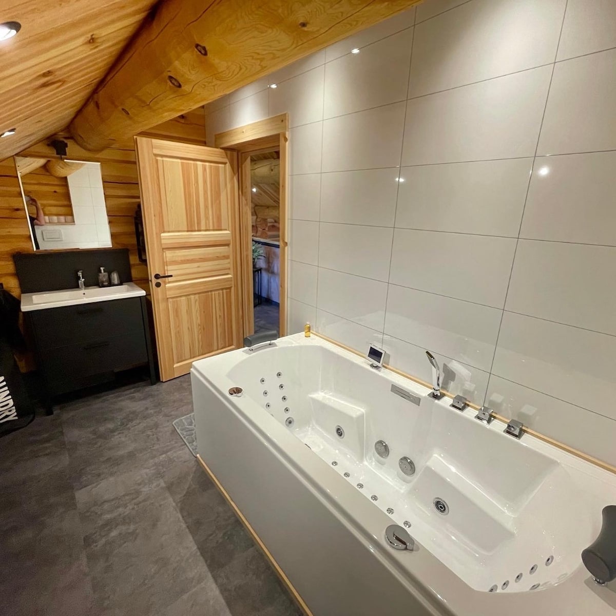A spacious bathroom features a luxurious two-person whirlpool tub, elegantly designed with modern fixtures. The walls showcase sleek tiles, complemented by natural wooden accents. A dark cabinet stands beside a contemporary sink, providing functional convenience. Soft lighting enhances the room's inviting atmosphere.