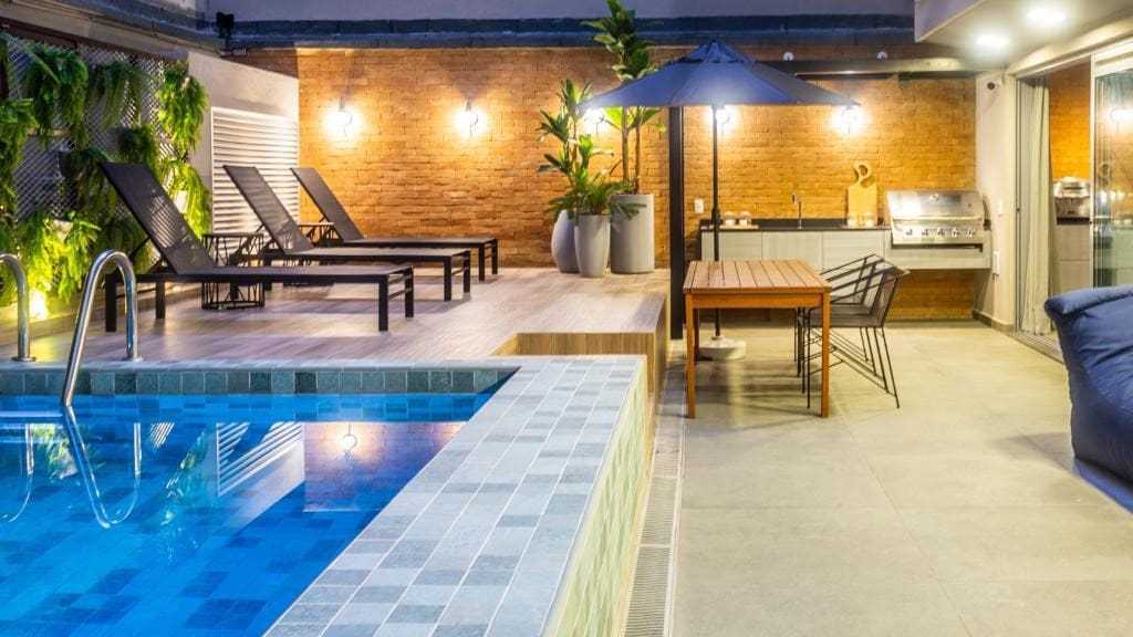 An outdoor space features a refreshing pool with a tiled edge, surrounded by sun loungers. A dining area is equipped with a wooden table and chairs, while a barbecue grill is positioned nearby. Lush greenery and a large umbrella add to the inviting atmosphere.