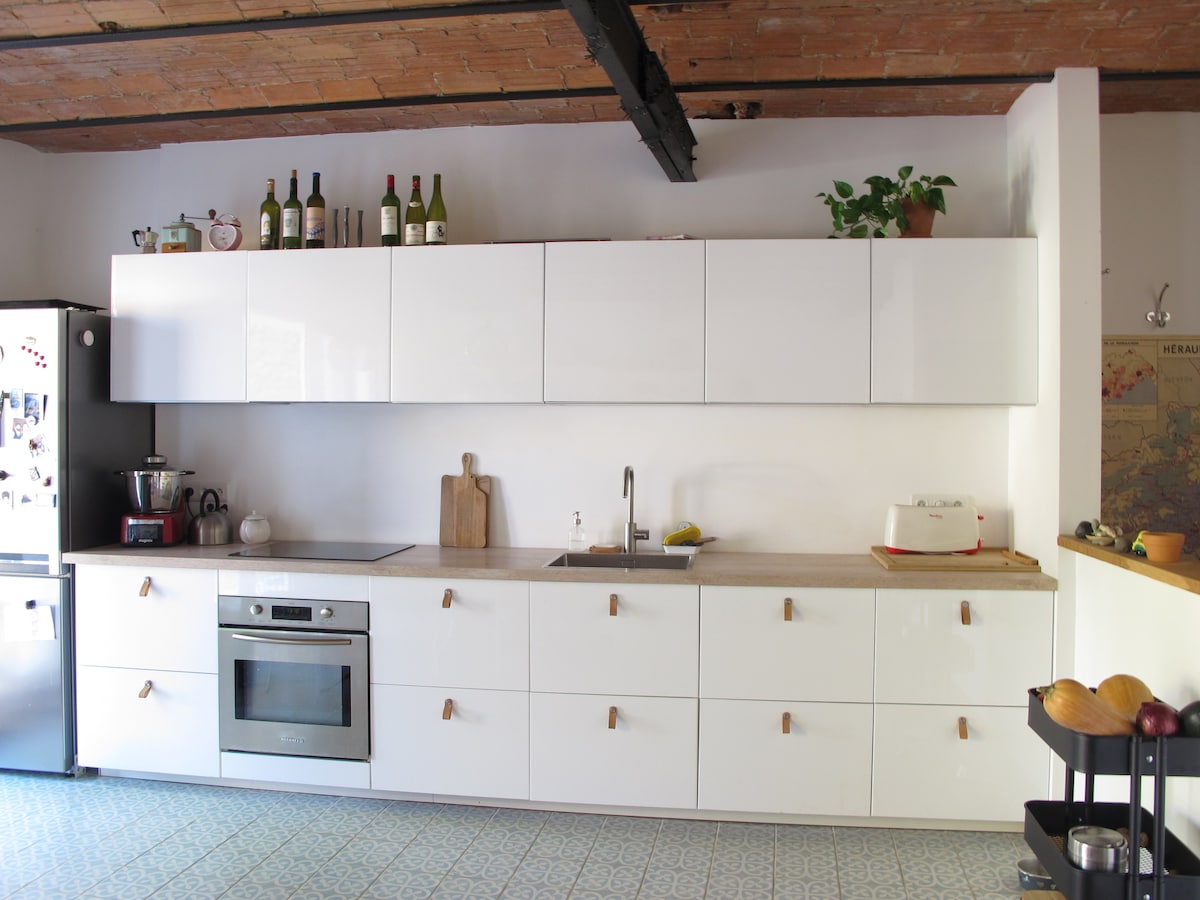 A modern kitchen space features sleek white cabinetry with natural wood handles, providing a clean aesthetic. Stainless steel appliances, including a built-in oven, are neatly integrated. A sink is positioned beneath an overhead cabinet, and decorative elements like wine bottles and a plant add a touch of character.