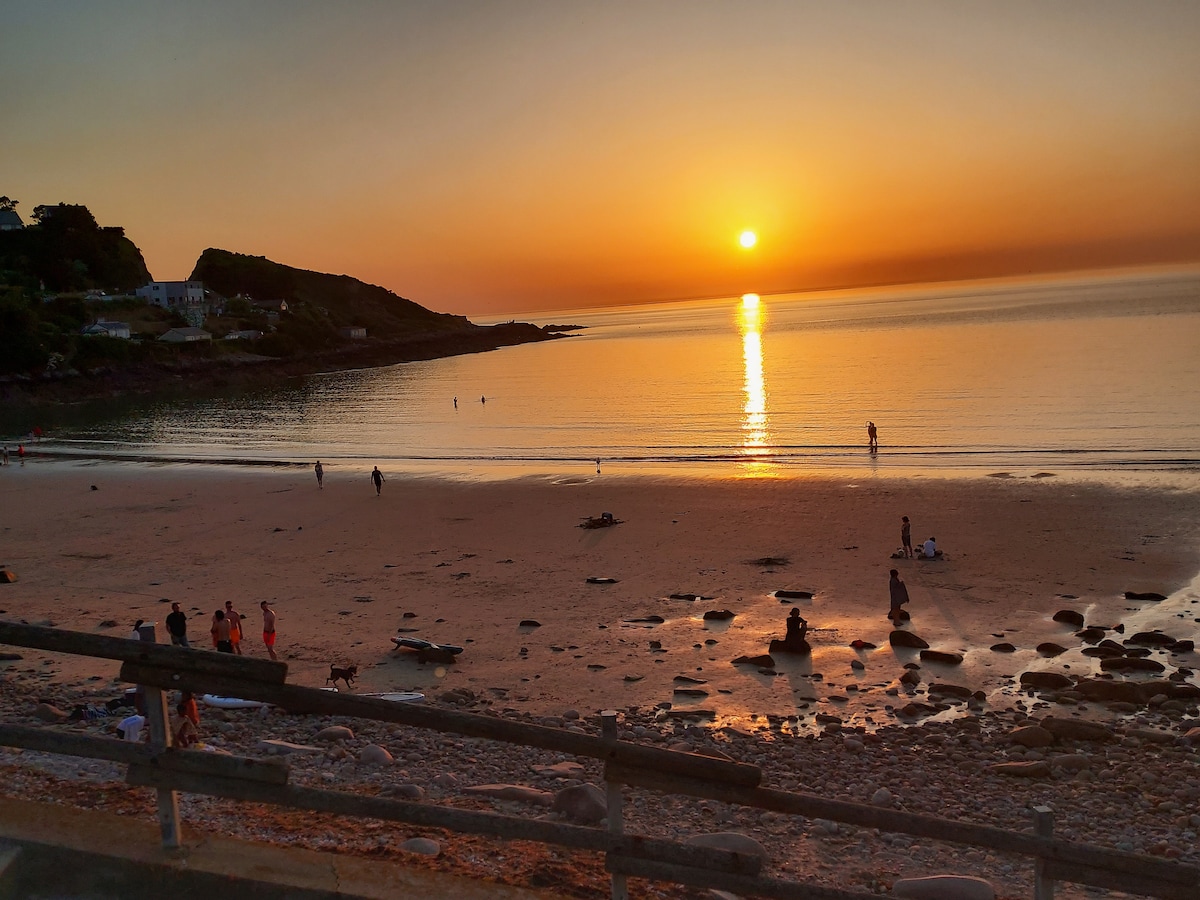 A serene sunset is captured over a calm beach, where the sun reflects on the gentle waves. Silhouettes of people can be seen enjoying the shoreline, and the shoreline features smooth stones, with a backdrop of cliffs and distant buildings.