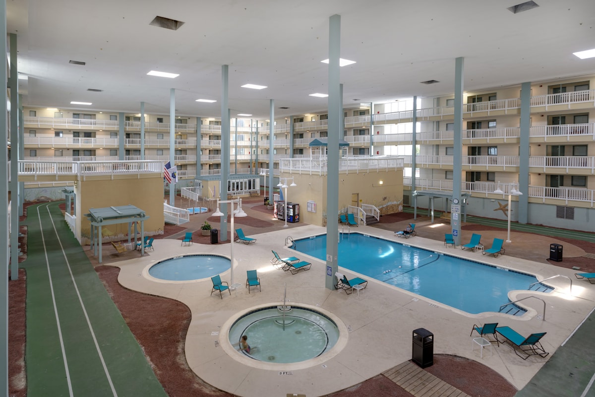 An indoor pool area features multiple swimming pools and hot tubs surrounded by lounge chairs in a spacious setting. Bright walls and natural light fill the space, while an indoor running track is visible along one edge, enhancing the recreational options available.