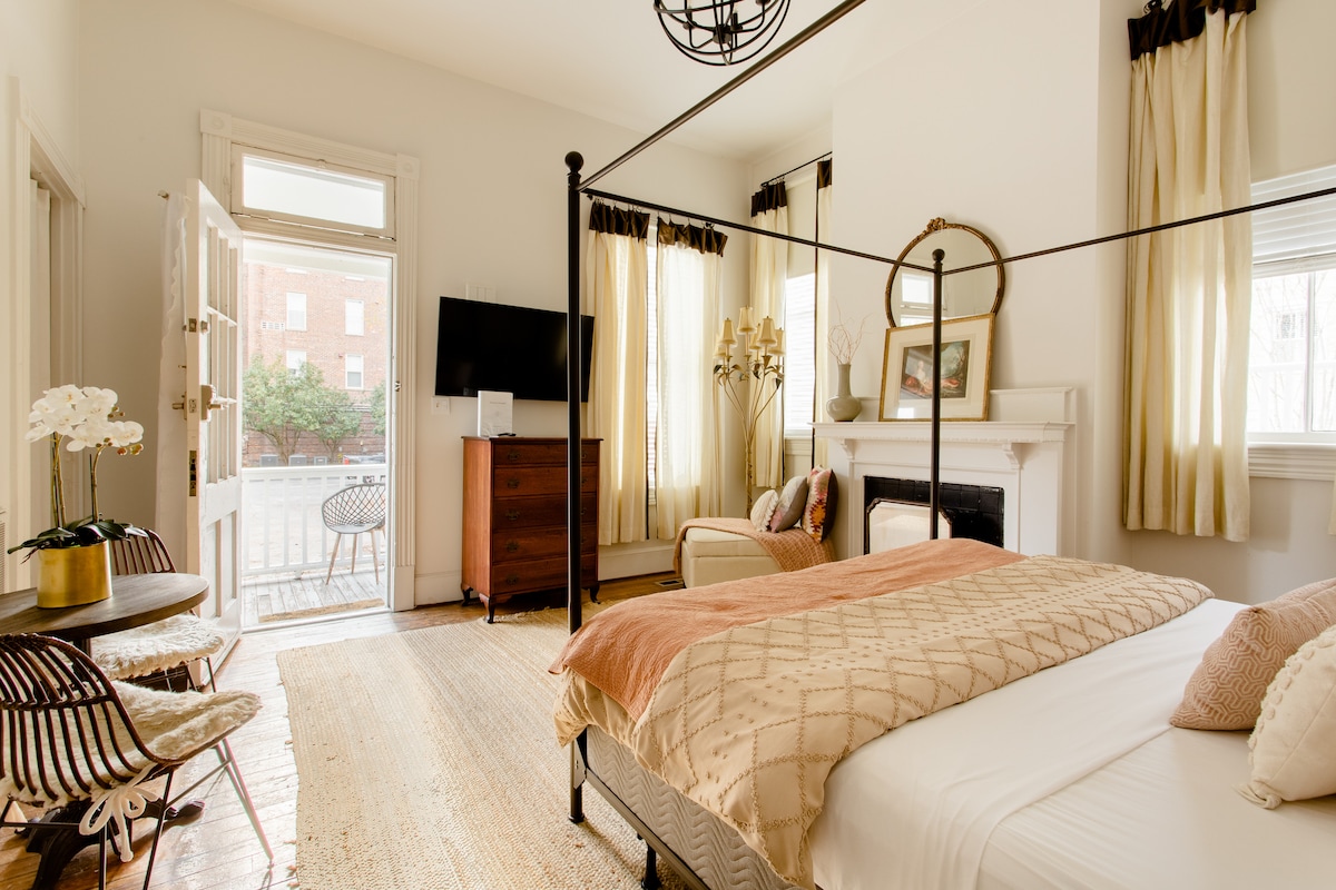 The studio apartment features a luxurious king-size canopy bed with a light-colored quilt. Natural light enters through spacious windows adorned with soft curtains. A cozy seating area offers modern furnishings, with a decorative fireplace and a small dining table completing the inviting space.