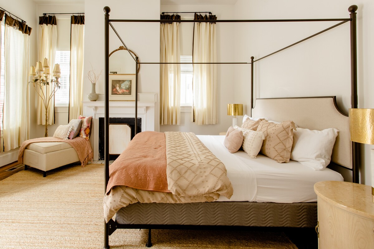 A well-appointed bedroom features a king-size canopy bed adorned with decorative pillows and a soft blanket. Light-colored curtains frame the windows, allowing natural light to enhance the space. A vintage fireplace serves as a focal point beside a cozy seating area with a chaise lounge.