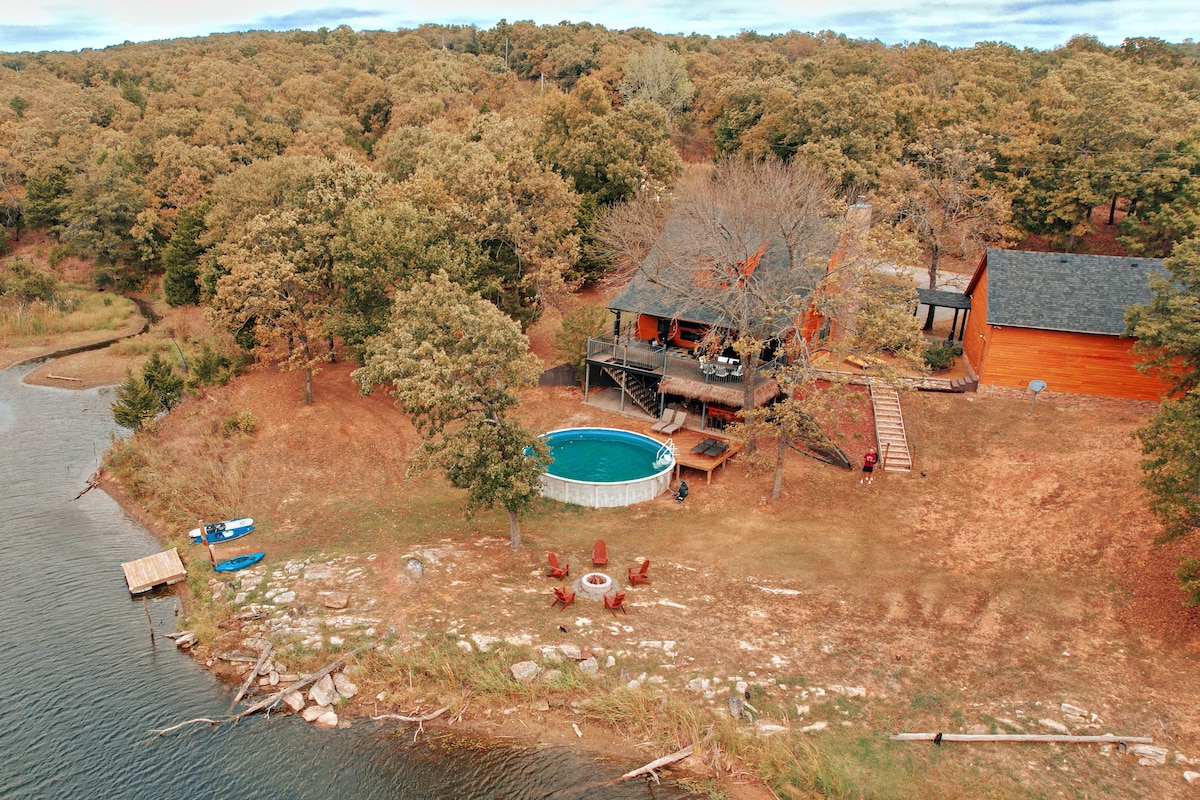 An aerial view showcases a cozy cabin nestled among trees, with a circular pool on a grassy area. A wooden deck extends from the cabin, leading to a firepit surrounded by chairs. Nearby, kayaks rest on the shoreline, offering easy water access.