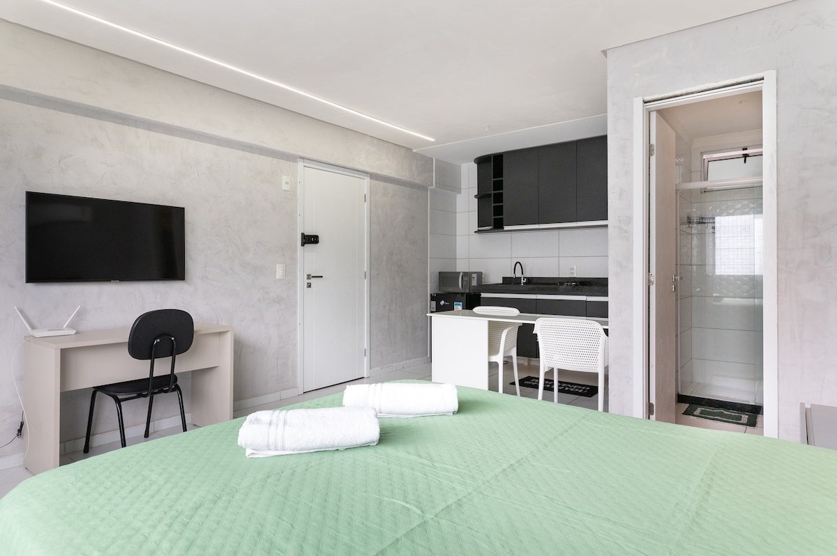 A bright interior showcasing a spacious room with a green bedspread covering a large bed. A modern kitchenette is visible, along with a compact dining area. A work desk with a chair is positioned nearby, and a doorway leads to a contemporary bathroom featuring a glass shower.