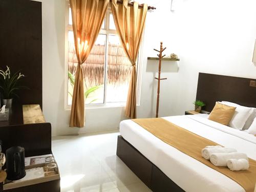 A well-appointed bedroom features a spacious bed with a bolster and neatly arranged towels. Natural light filters through large windows adorned with soft curtains. A minimalist decor is complemented by a wooden dresser and a small shelf, contributing to a serene ambiance.