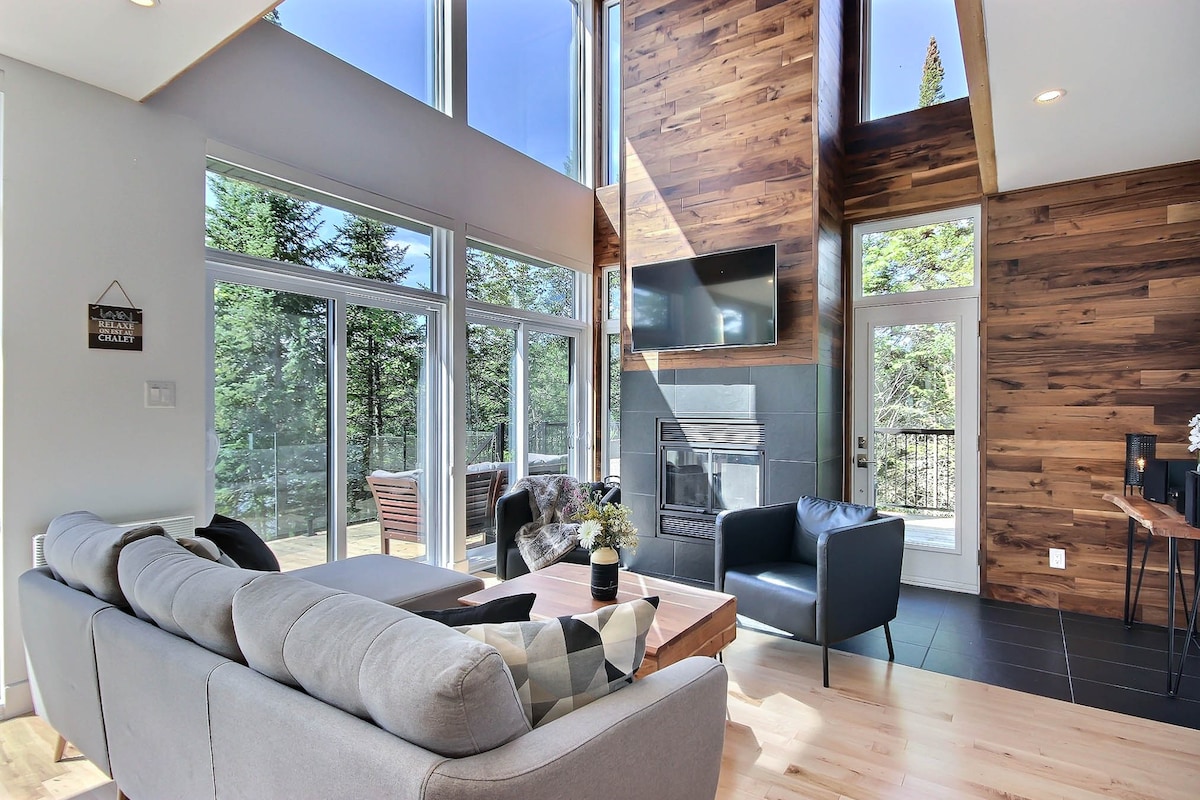 A bright living area features tall windows allowing natural light to illuminate the space. A comfortable sofa and armchairs are positioned facing a wall-mounted television and a modern fireplace. The wood paneling on the walls adds warmth to the contemporary design.