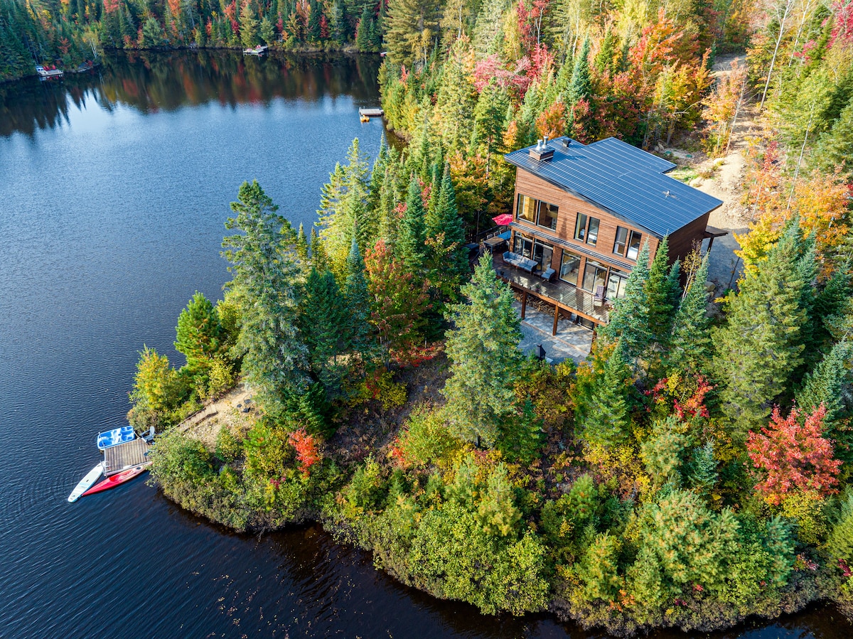 An aerial view captures the chalet nestled among vibrant autumn foliage alongside a tranquil lake. The two-story structure features a deck that extends into the greenery, while a private dock is visible by the water's edge, complementing the natural surroundings.
