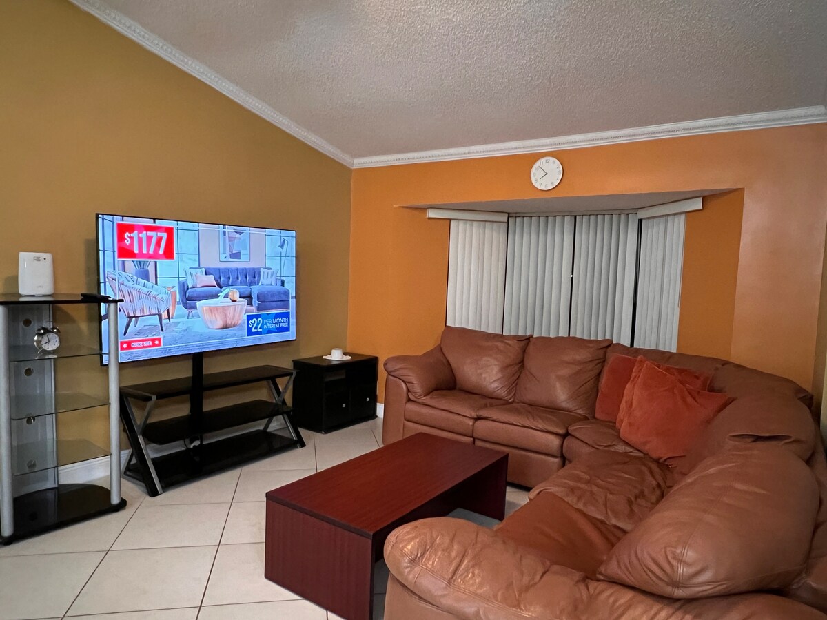A spacious living area features a large, brown sectional sofa with decorative pillows. A coffee table sits in the center, while a flat-screen TV displays a colorful image. Light streams in through vertical blinds, illuminating the room's warm orange walls.