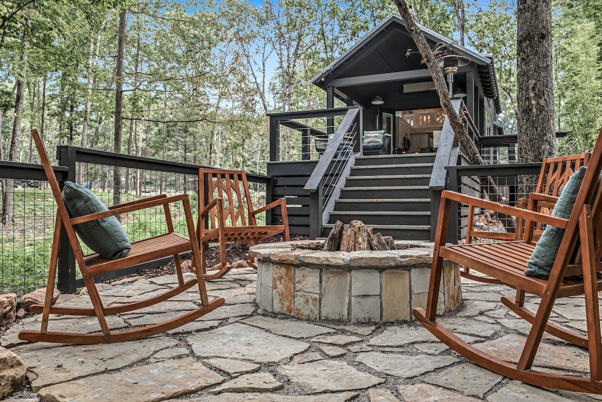 A stone fire pit is surrounded by four wooden rocking chairs placed on a natural stone patio. The tiny home is visible in the background, set among trees that provide a serene atmosphere. Stairs lead up to the entrance of the home.