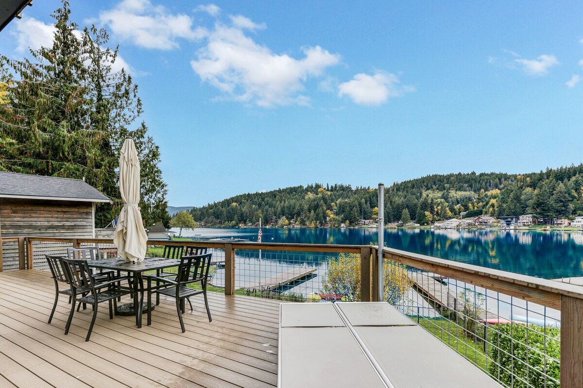 A spacious deck features a large table surrounded by black metal chairs, offering views of the serene lake and surrounding greenery. The calm water reflects the blue sky and trees, creating a peaceful atmosphere for relaxation and outdoor dining.