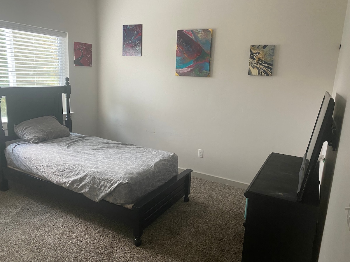 A minimalist room featuring a single bed with a grey patterned bedding set. Four abstract art pieces are displayed on light beige walls. A television is positioned on a dark dresser, and natural light pours in through a window with blinds.