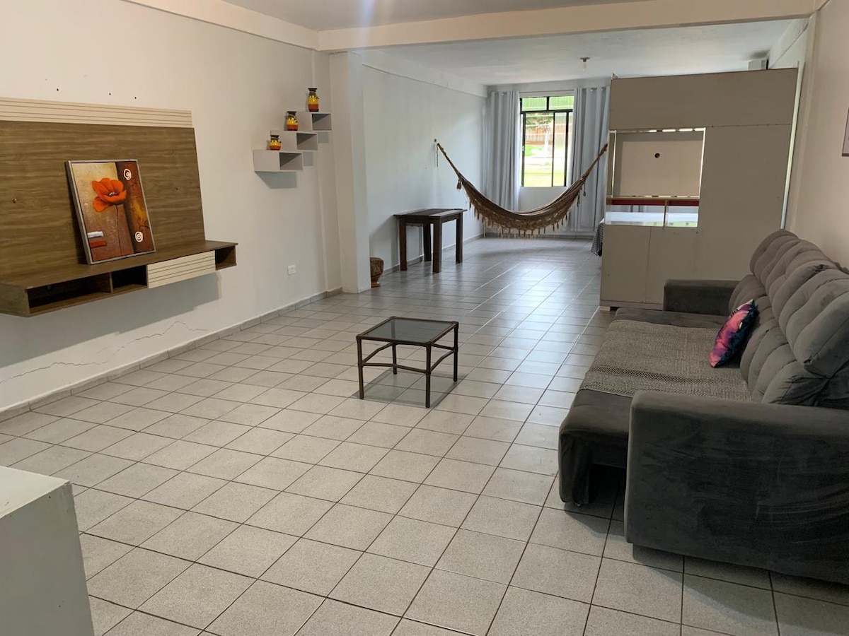 A spacious living area is featured, with neutral tiled flooring and ample natural light. A comfortable grey sofa is situated near a low coffee table. Decorative shelves are mounted on the wall, and a hammock is seen in the background, adding to the inviting and relaxed atmosphere.