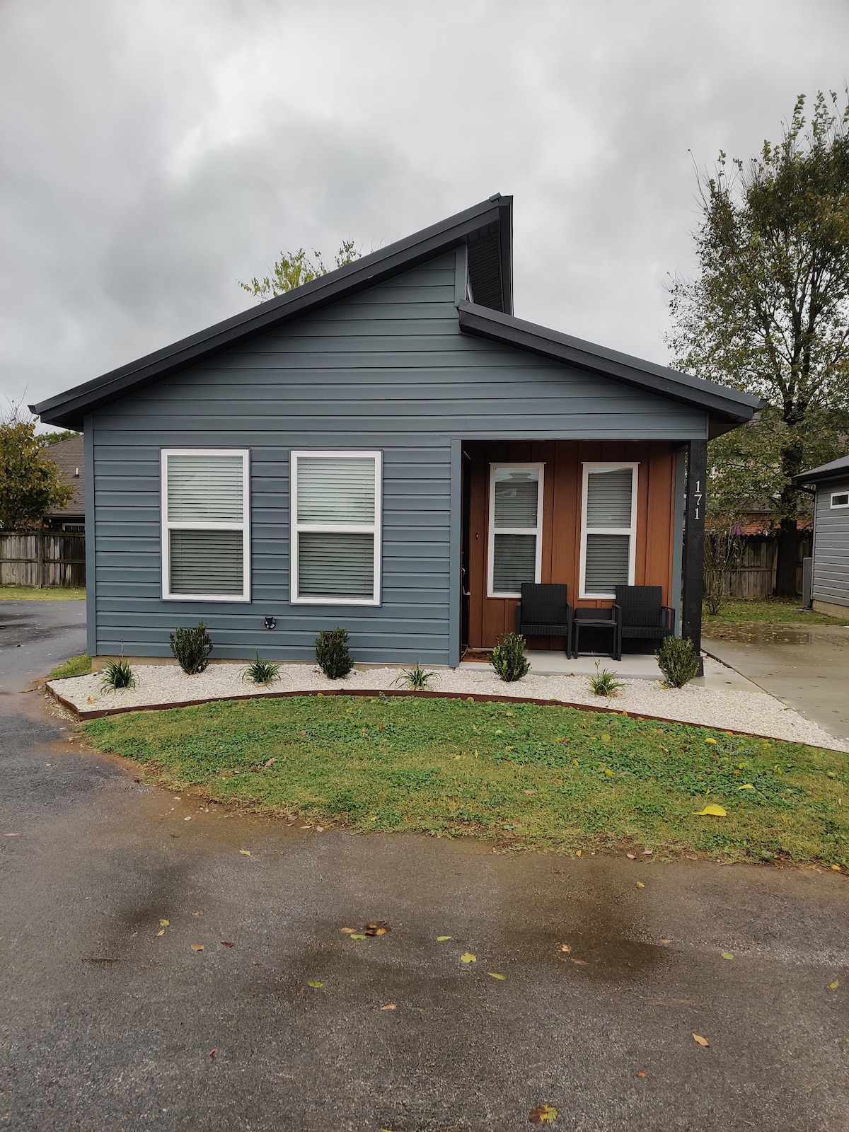 A modern tiny home features a distinctive roofline and a blue exterior. Two large windows allow natural light to enter, while a small porch with two chairs invites relaxation. Neatly designed landscaping with low plants enhances the home’s curb appeal.