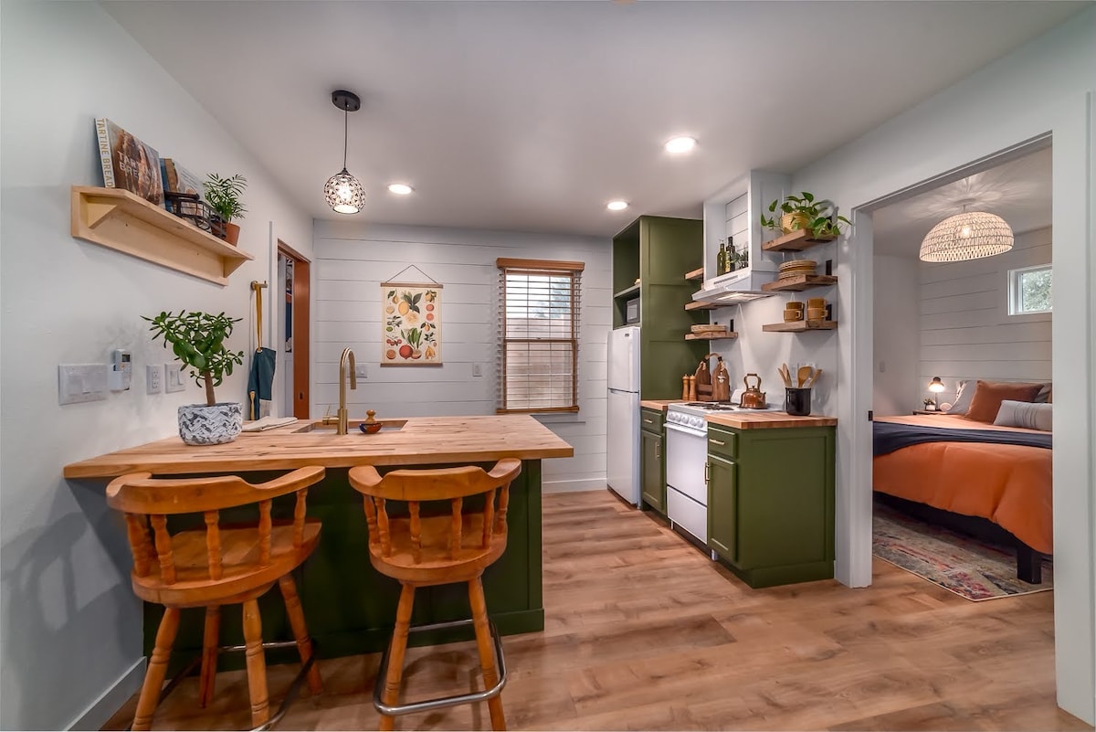 A well-appointed kitchen area features a wooden bar with two stools beside olive green cabinetry. Shelf space holds plants and kitchenware, while a bright window allows natural light. The adjoining room partially reveals a cozy sleeping area in soft tones.