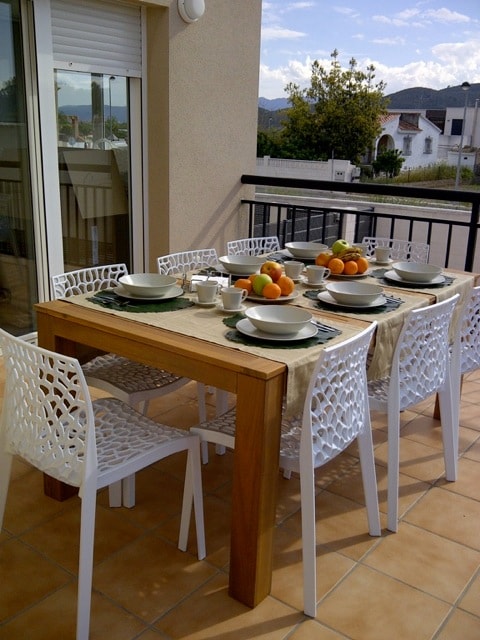 An outdoor dining area is presented, featuring a wooden table surrounded by six white chairs. The table is set with dishes, cups, and a centerpiece of oranges and other fruits. A balcony railing provides a view of the surroundings, with sunlight illuminating the space.