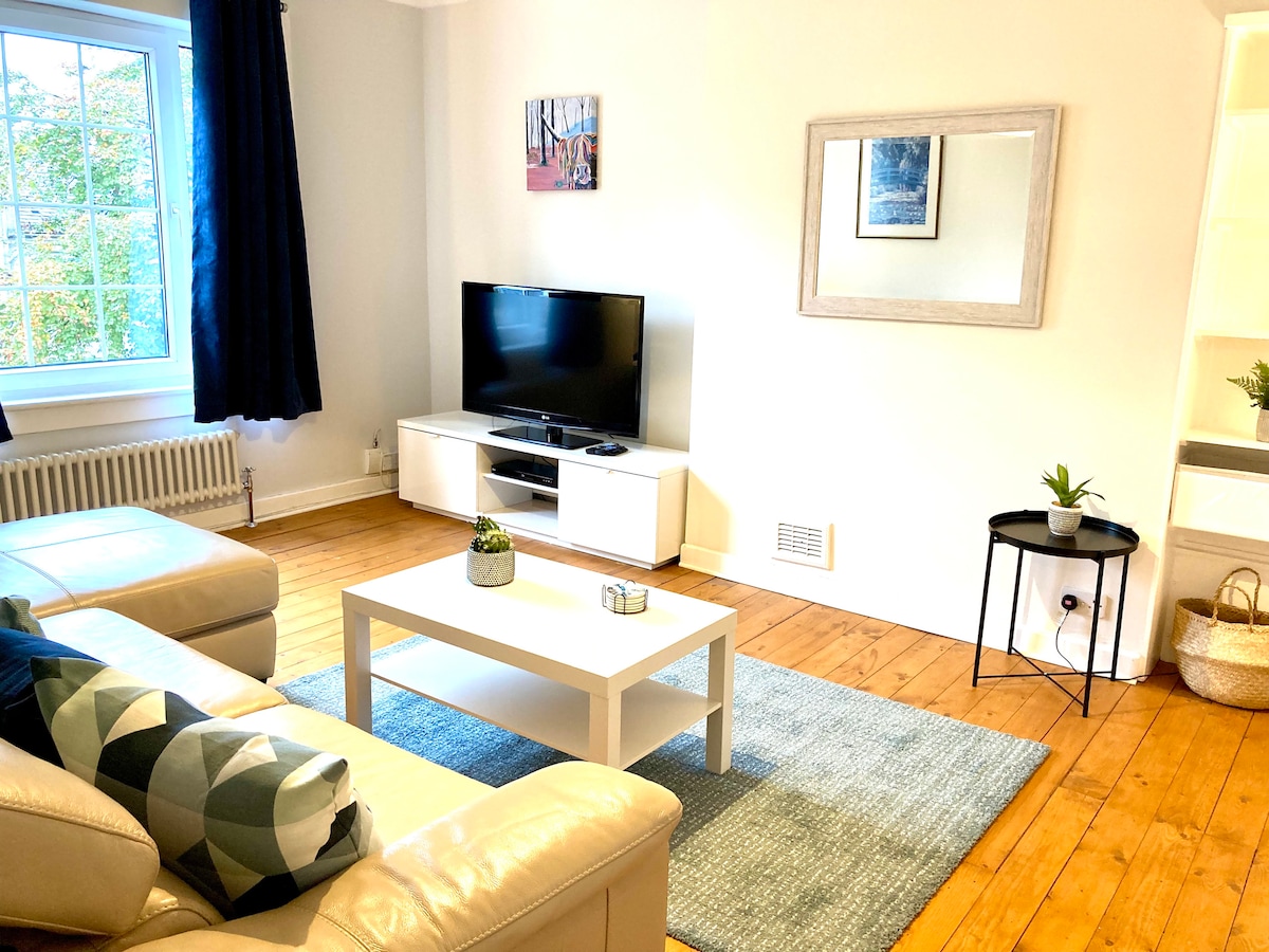 A comfortable lounge is presented with a sectional leather sofa and a coffee table atop a soft area rug. A flat-screen television is positioned against a light wall, complemented by a large window with dark curtains, allowing natural light to brighten the space.