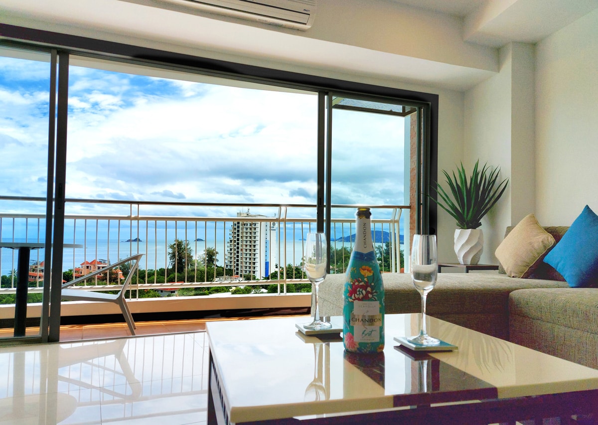 A modern lounge area features a comfortable sofa and a glass coffee table, accompanied by two glasses and a bottle of water. Floor-to-ceiling windows open to reveal stunning views of the coastline and sea, while a potted plant adds a touch of greenery.