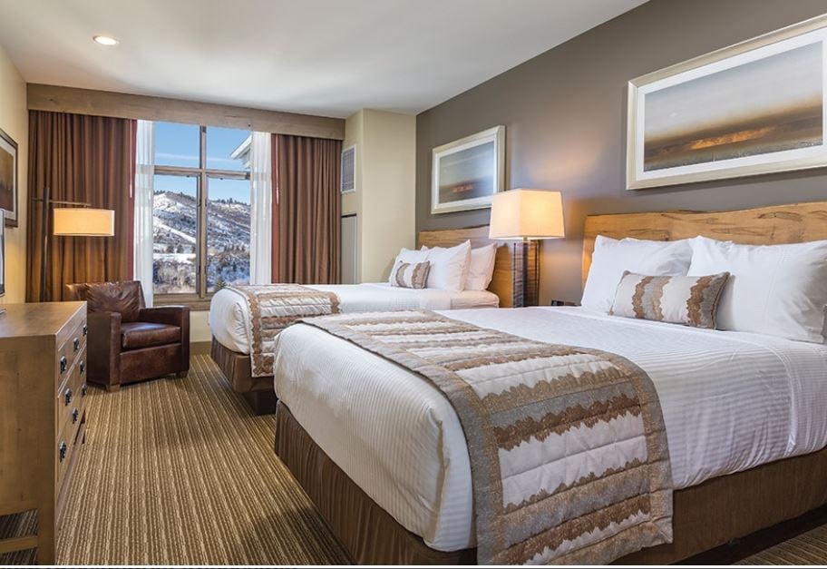 A spacious hotel room contains two large beds with neutral-toned linens and decorative pillows. A wooden dresser and a dark leather armchair are positioned near a large window, offering a view of the mountains. Soft lighting creates a calming atmosphere.