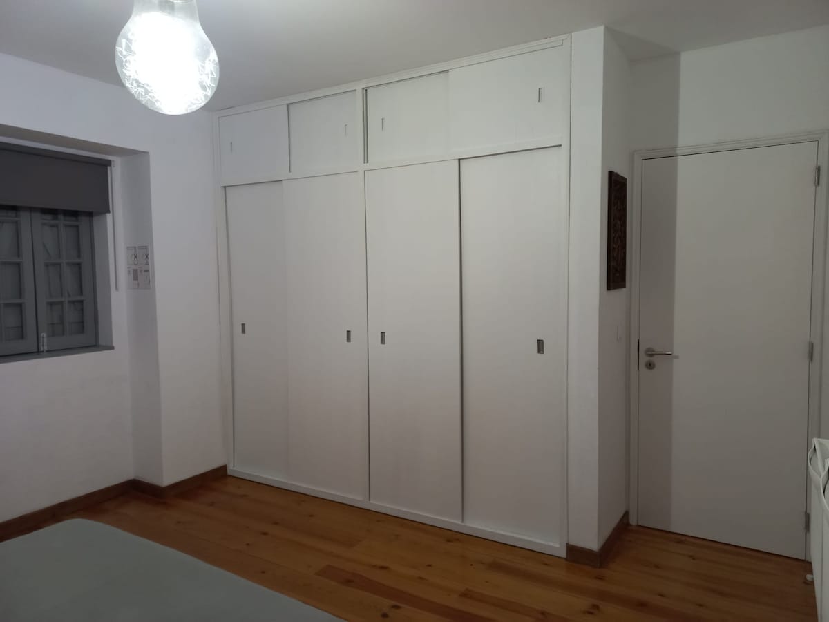 A well-organized space featuring built-in white wardrobes against a light-colored wall. The wooden floor adds warmth to the room, and a simple door leads to another area. Natural light filters in through a window with a grey blind.