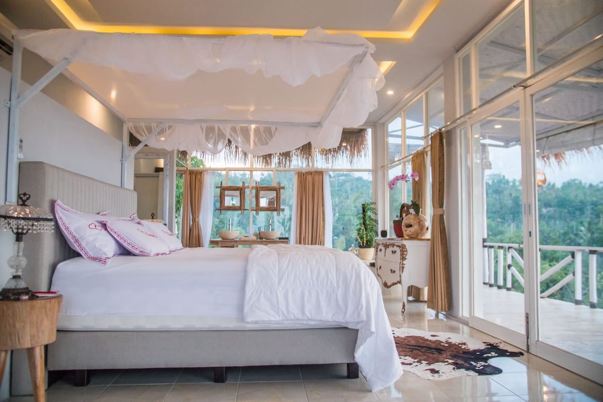 A spacious bedroom features a four-poster bed draped with light fabric, complemented by a wooden nightstand. Expansive glass doors provide access to a scenic view, while elegant furnishings enhance the serene ambiance. Natural light fills the room, reflecting off the tiled floor.