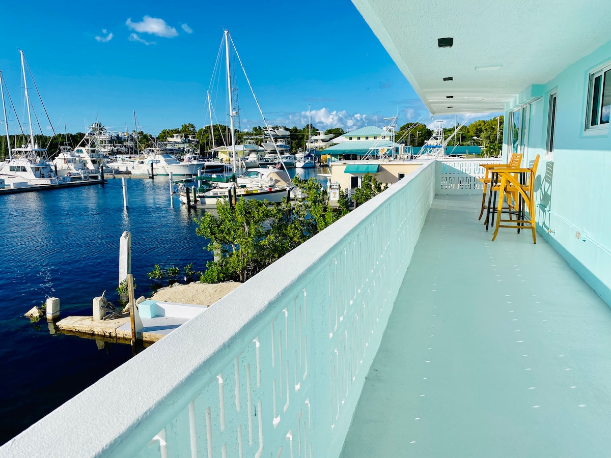 A wide balcony overlooks a serene harbor, featuring several boats docked in the water. Two tall chairs are positioned against the railing, offering a view of the vibrant blue sky and surrounding marina.