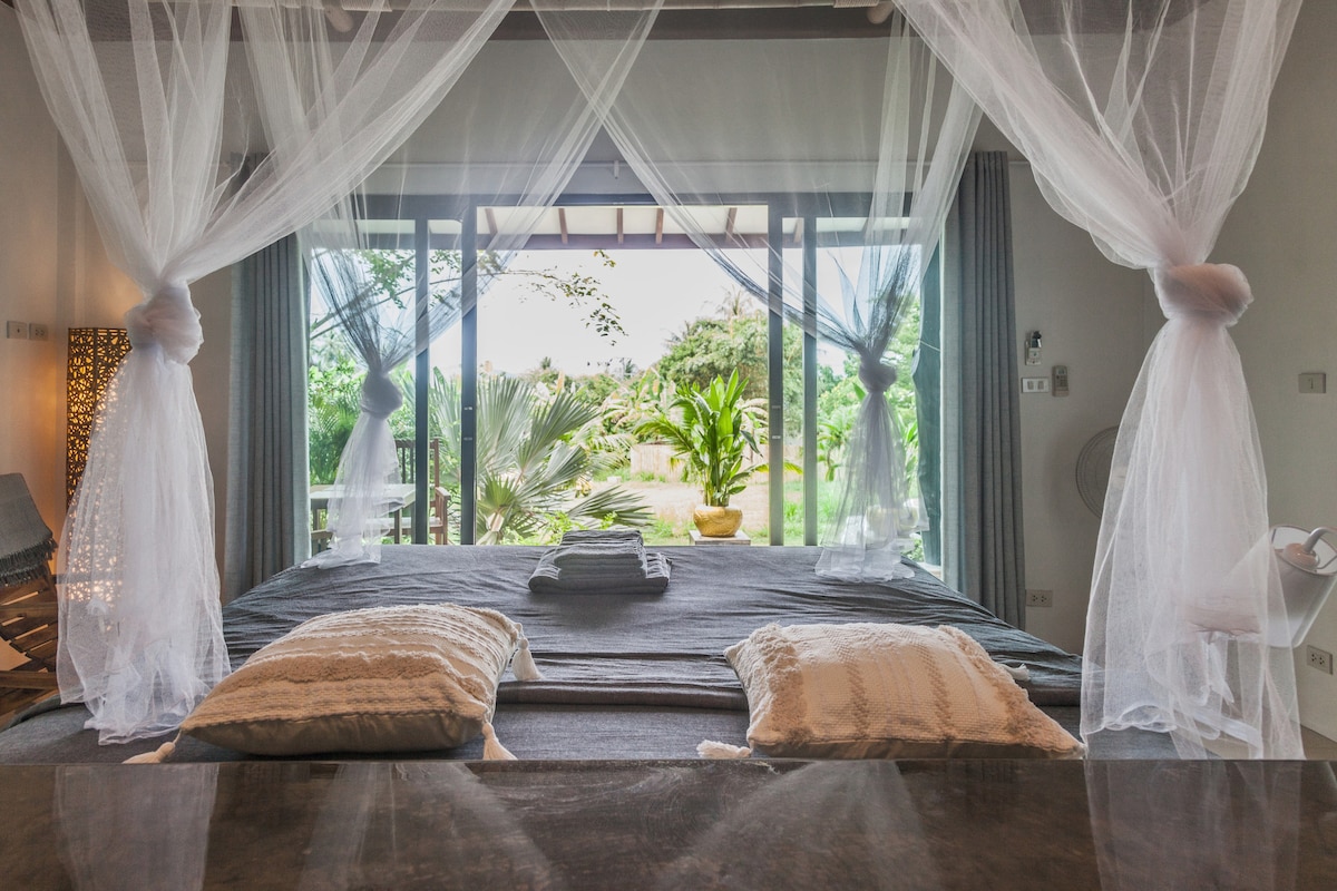 A cozy sleeping area features a spacious bed draped with sheer curtains, allowing natural light to illuminate the room. Two decorative pillows rest at the foot of the bed, while sliding glass doors open to a view of lush greenery outside.