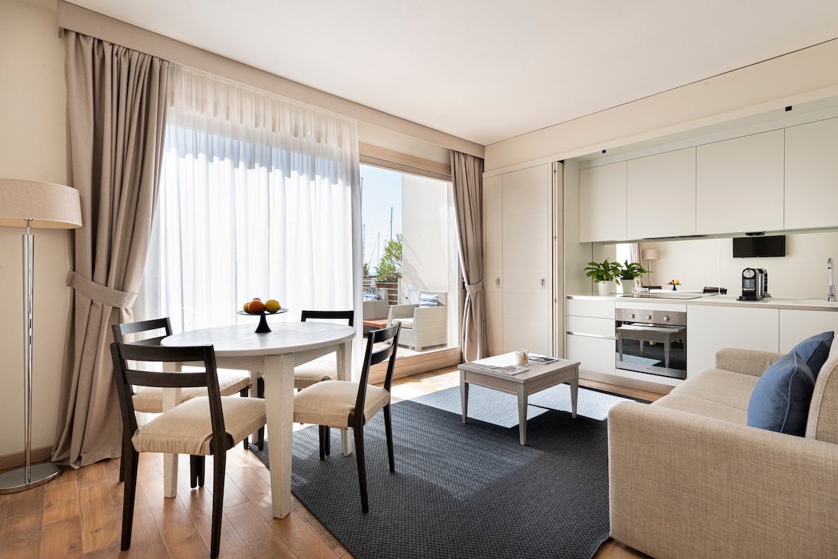 A bright living area is presented featuring a round dining table with four chairs, a comfortable sofa, and a modern kitchenette. Large windows allow natural light to fill the space, which is complemented by light wood flooring and neutral-colored furnishings.