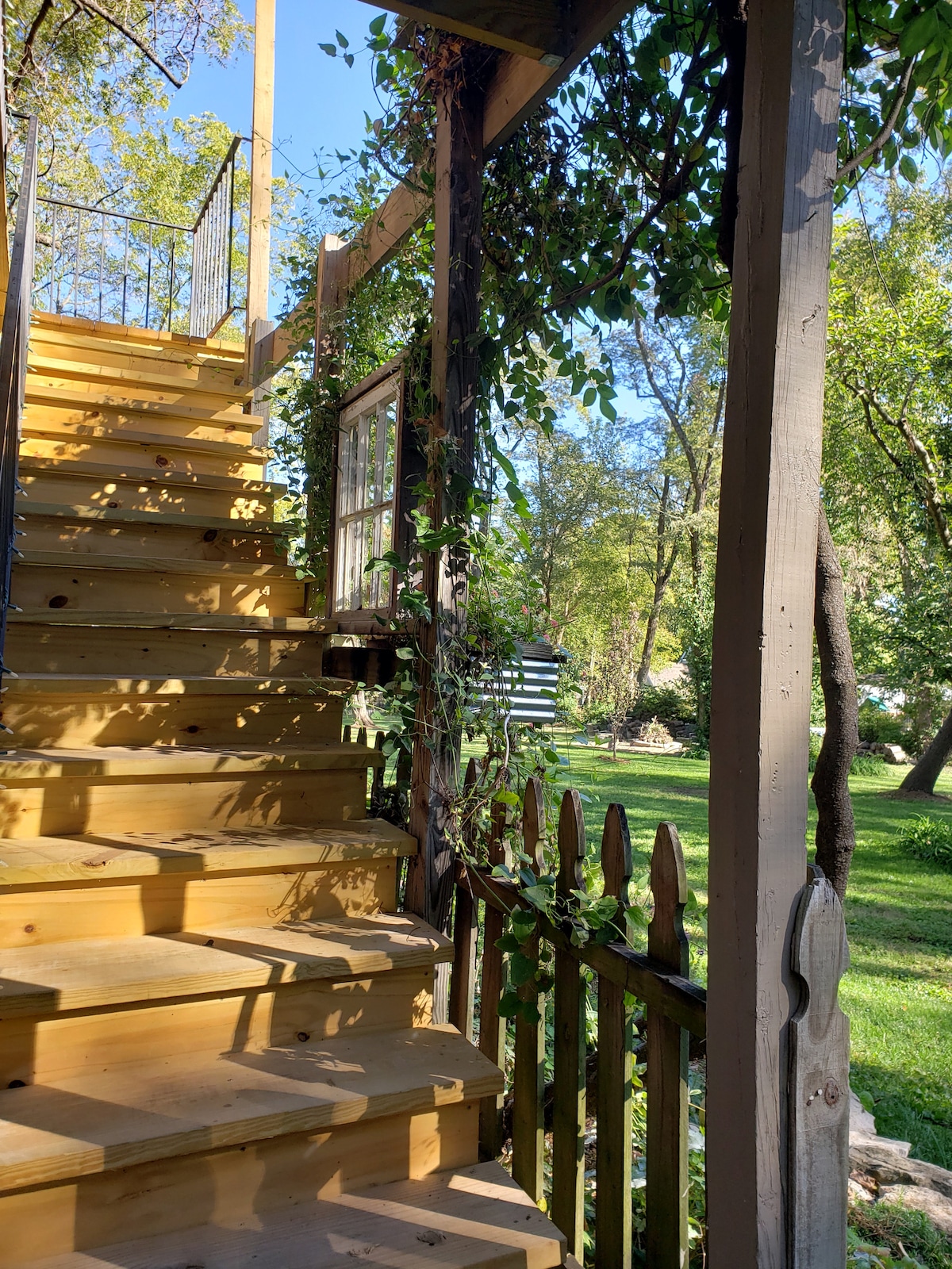 Wooden stairs ascend through lush greenery, adorned with a railing and framed by a rustic wooden fence. Sunlight filters through the leaves, illuminating the path to the second story of the structure.