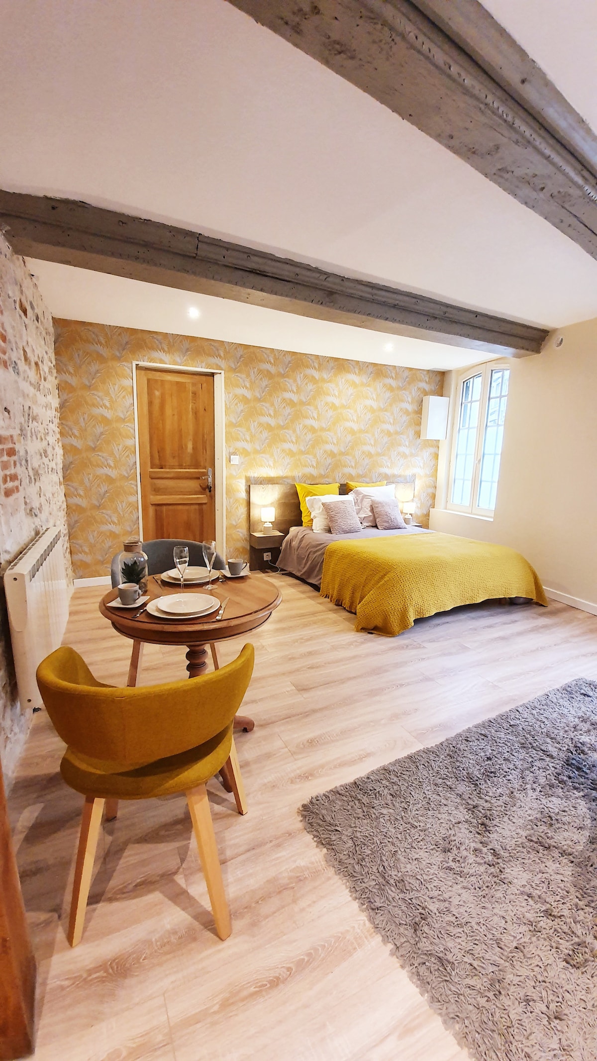 A spacious living area is featured, with a double bed dressed in warm textiles and illuminated by bedside lamps. A small dining table is set for two, complemented by a yellow chair. The textured wall and wooden beams enhance the character of the room.