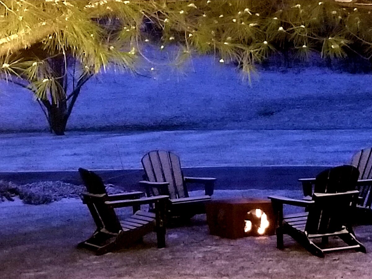 A cozy outdoor area features four heavy-duty Adirondack chairs surrounding a firepit, with firewood nearby for use. Soft outdoor string lighting enhances the ambiance, and a peaceful park-like landscape is visible in the background, creating a serene setting for gatherings.