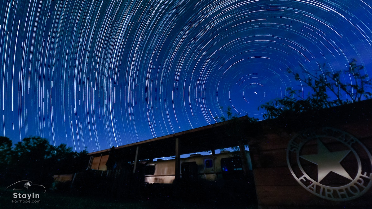 A night sky is filled with star trails, creating a dynamic display of movement. A silhouetted structure is visible in the foreground, with a covered area extending alongside, emphasizing the peaceful rural surroundings.