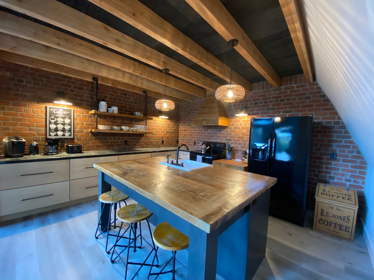 The kitchen features a modern design with exposed brick walls and wooden beams overhead. A large island with a wooden countertop is surrounded by three stools. Pendant lights hang above the island, illuminating various kitchen appliances and shelving filled with dishes.