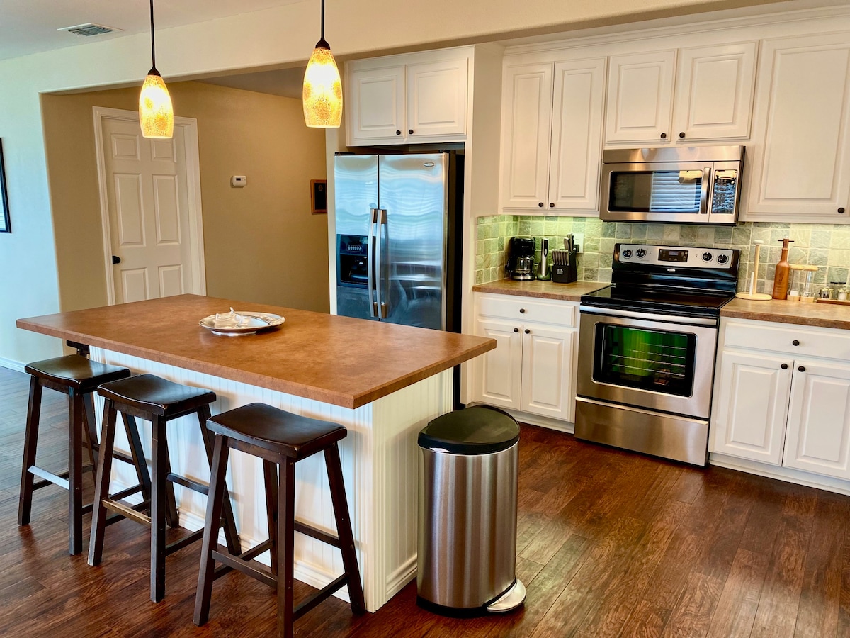 An open concept kitchen is featured with a central island and three wooden bar stools. Stainless steel appliances, including a refrigerator and oven, are complemented by white cabinetry and a tiled backsplash. Pendant lights hang above the island, and warm wooden flooring completes the space.
