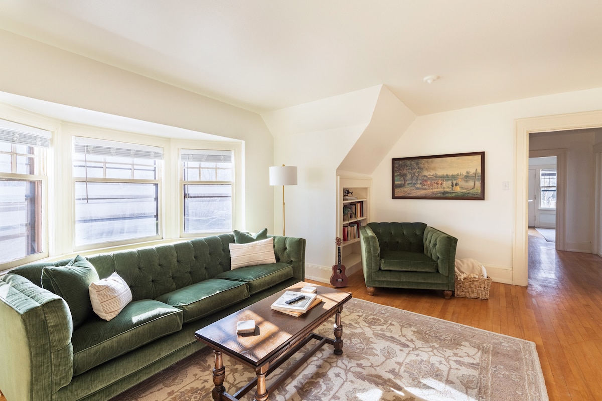 A comfortable living room features olive green upholstery on a sofa and an accent chair. A wooden coffee table sits on a light-colored area rug, complemented by natural light filtering through multiple windows. Wall art and a lamp add functional and decorative elements.