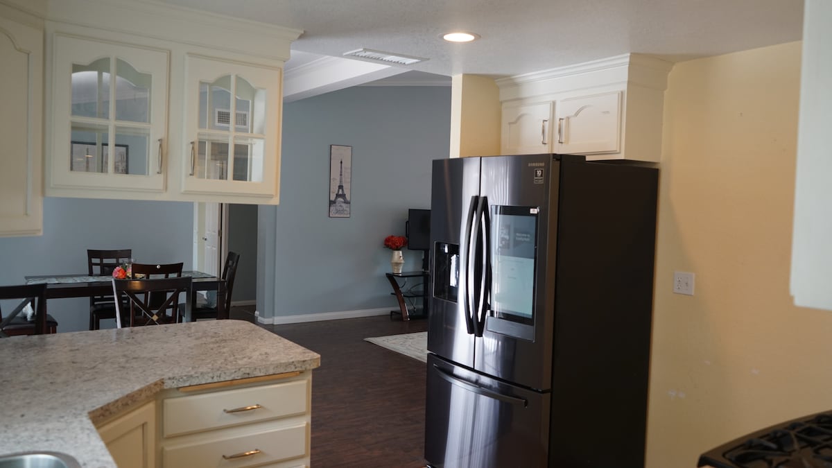 A modern kitchen is visible, featuring light-colored cabinetry with glass-fronted doors. A spacious granite countertop is paired with a black refrigerator, while a dining area in the background is indicated by wooden chairs and a table. Soft natural light brightens the space.