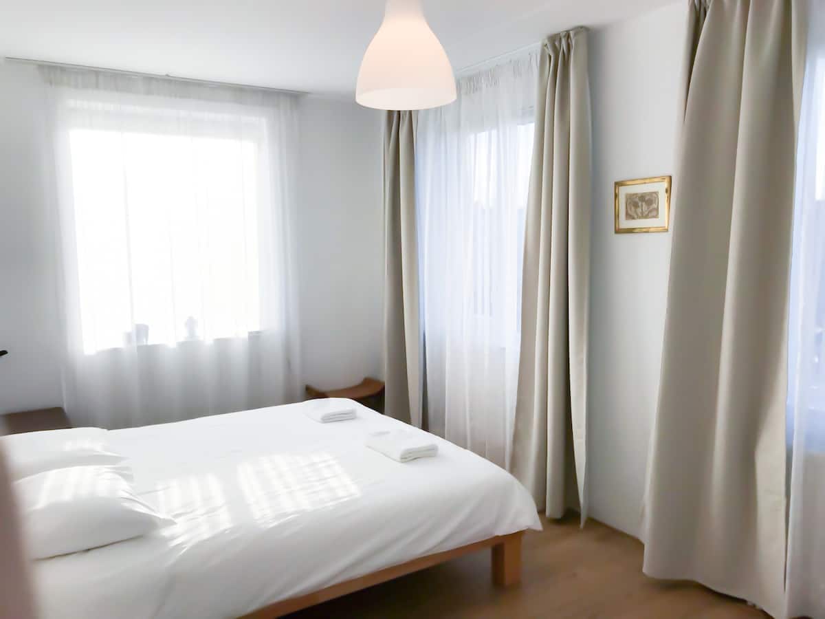 A bright bedroom features a spacious bed with crisp white linens and two neatly placed towels. Large windows are dressed with sheer curtains that allow soft natural light to fill the room. A simple piece of framed art hangs on the wall, contributing to the serene ambiance.