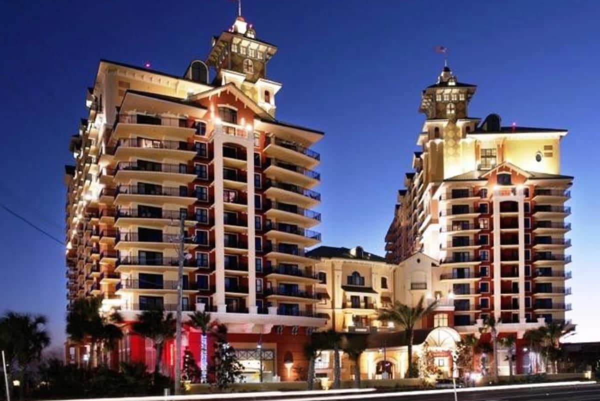 The exterior of a multi-story building is illuminated at night, showcasing unique architecture with twin towers featuring rooftop elements. Palm trees are visible in the foreground, and the entrance area is well-lit, enhancing the property's welcoming presence.