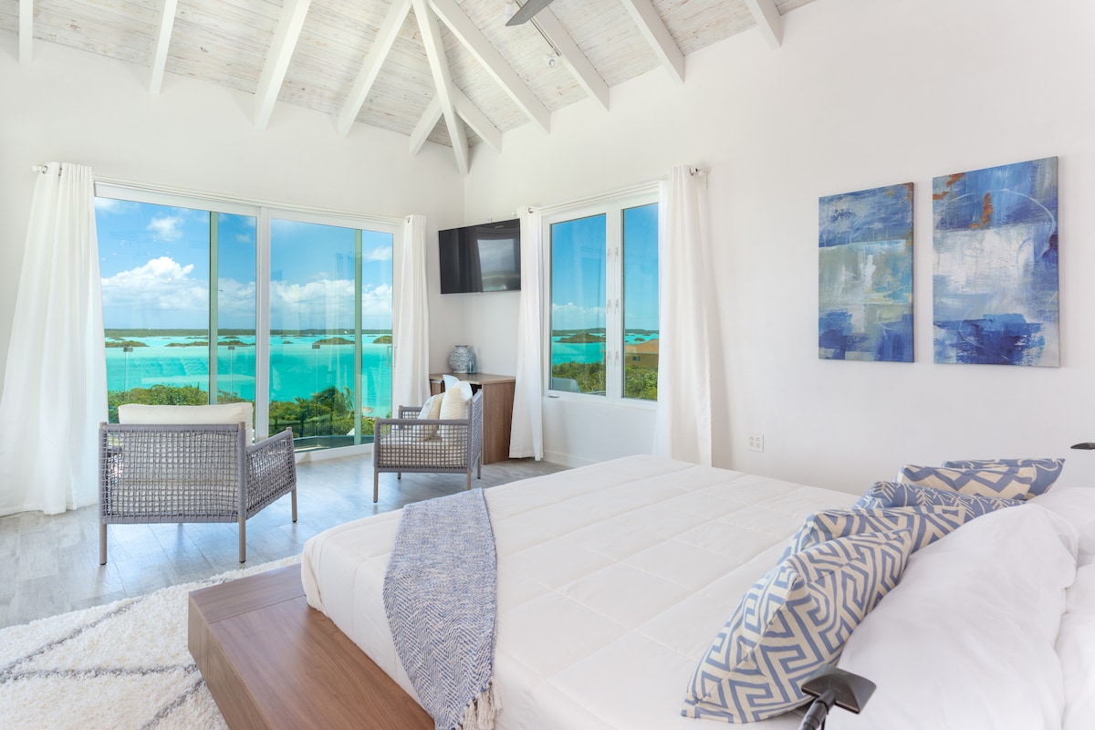 A master bedroom showcases large windows offering panoramic views of turquoise waters. The room features a spacious bed complemented by soft linens, while a cozy seating area with modern furniture enhances the inviting space. Light beams in through sheer curtains, creating a bright atmosphere.