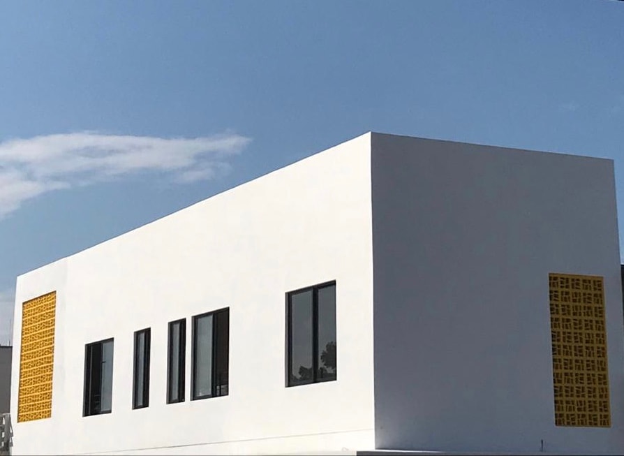 A modern white structure is displayed under a clear blue sky, featuring large black-framed windows that provide ample natural light. Yellow decorative panels are positioned symmetrically on the sides, adding a distinctive architectural detail to the building's exterior.