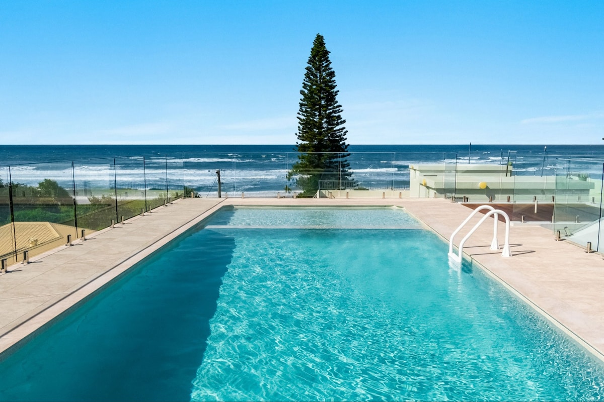 An outdoor pool area features a large swimming pool with clear blue water and a shallow section. Glass railings provide unobstructed views of the ocean and beach, while a tall pine tree stands in the background beneath a clear blue sky.