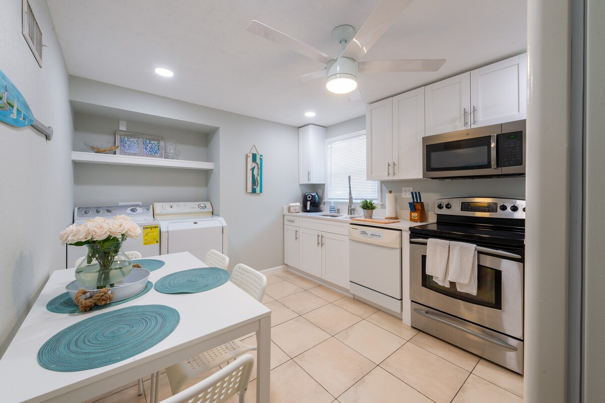 A modern kitchen space is equipped with stainless steel appliances, including a microwave and oven. A dining table, set with textured teal placemats, accommodates four. Open shelving displays decorative items, while a laundry area is conveniently integrated into the design. A ceiling fan enhances air circulation.