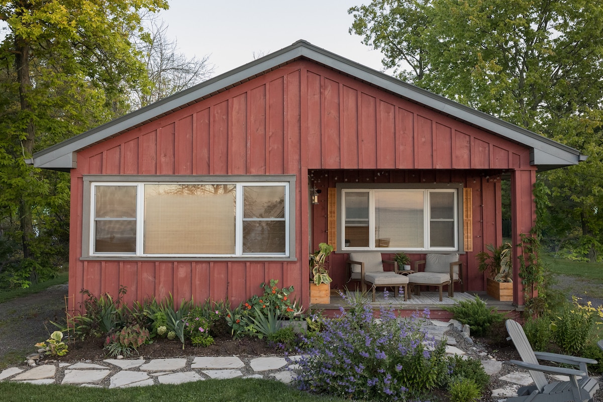 A charming red exterior defines the cottage, with large windows framing natural light and providing views of greenery. The front porch features comfortable seating surrounded by garden beds filled with flowering plants and neatly arranged stone pathways.