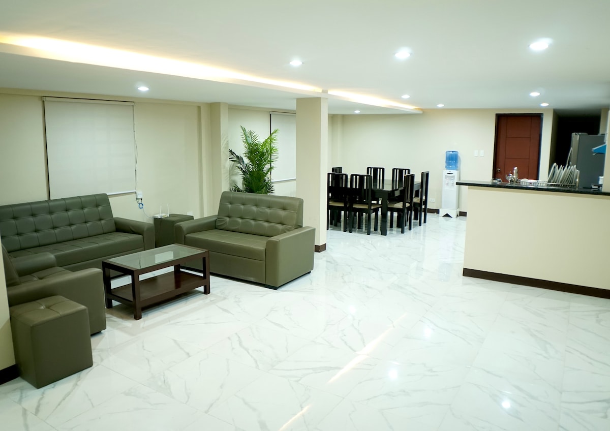 A modern living area features gray sofas arranged around a glass coffee table, with soft lighting enhancing the space. A dining area with a dark wooden table and several chairs is visible in the background, alongside a water dispenser.