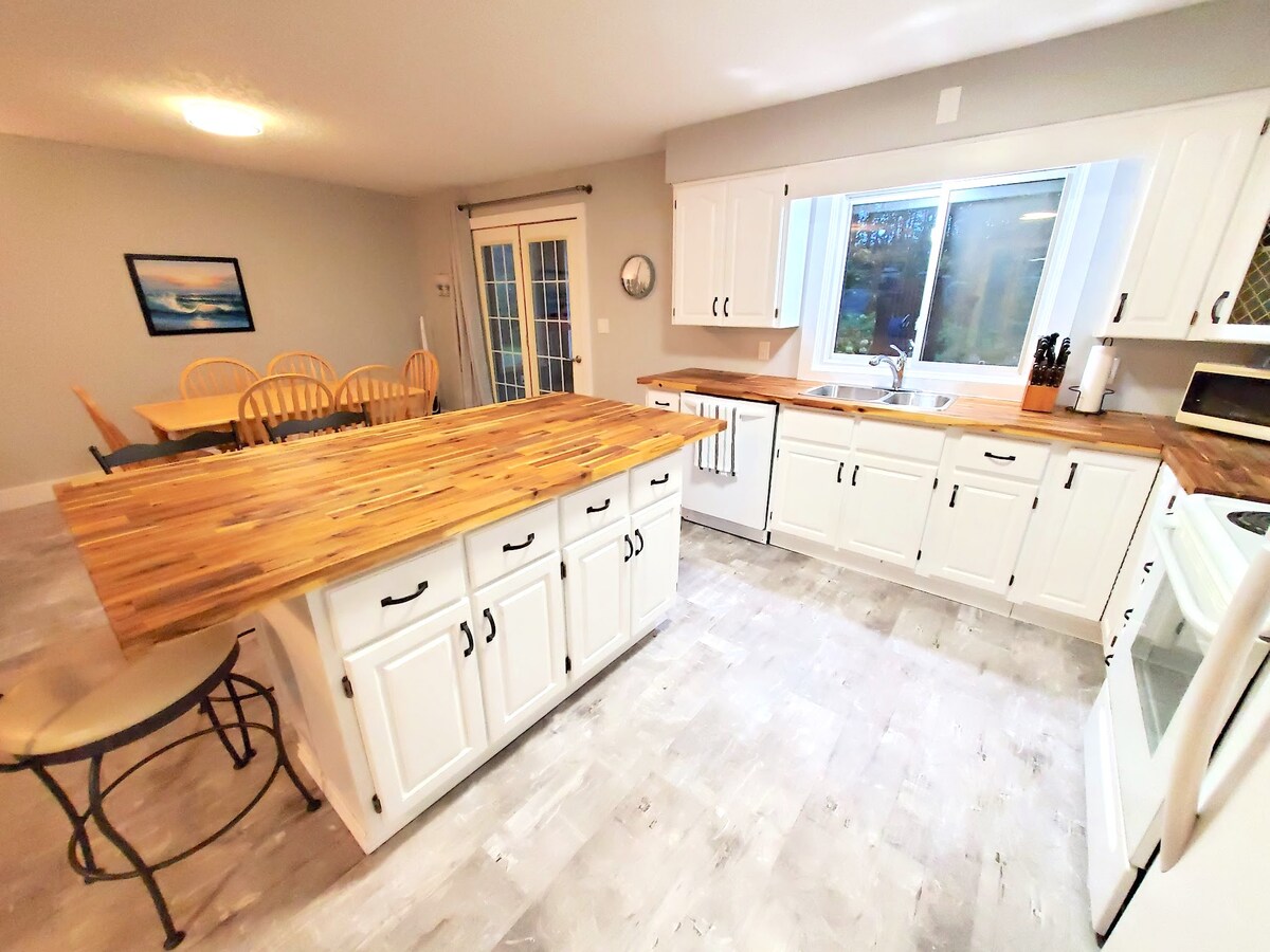 A modern kitchen is featured with white cabinetry and wooden countertops. A central island with seating is positioned beside a dining area, where a round table with six chairs is visible. Natural light enters through a large window, enhancing the spacious layout.
