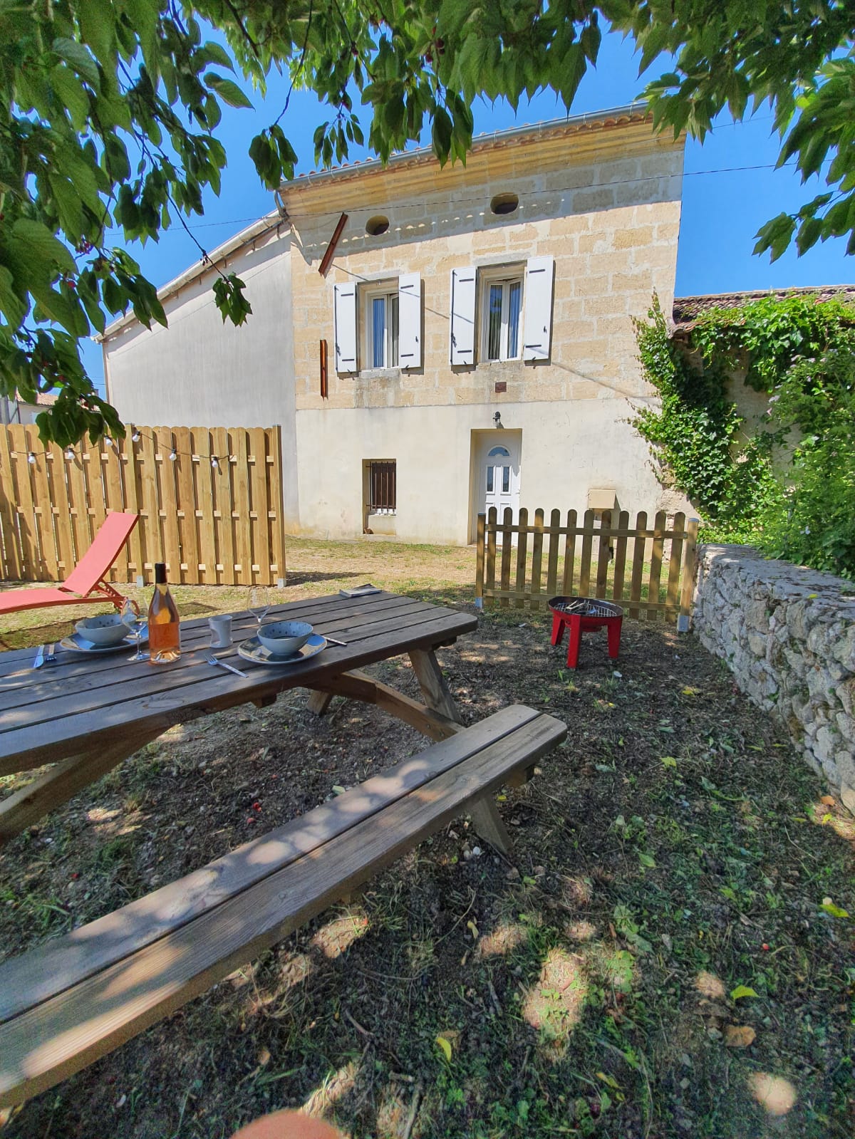 An outdoor seating area is set within a fenced garden, featuring a wooden picnic table with plates and a bottle on it. A red chair and a lounge chair provide additional seating options. The exterior wall of the house is visible in the background, surrounded by greenery.