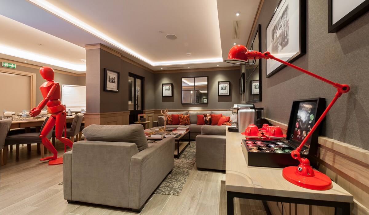 The common area features modern furnishings with neutral-toned sofas and a light wooden table. A bright red sculpture adds a pop of color, complemented by a bold red desk lamp. Framed artwork decorates the walls, and soft lighting enhances the ambiance.