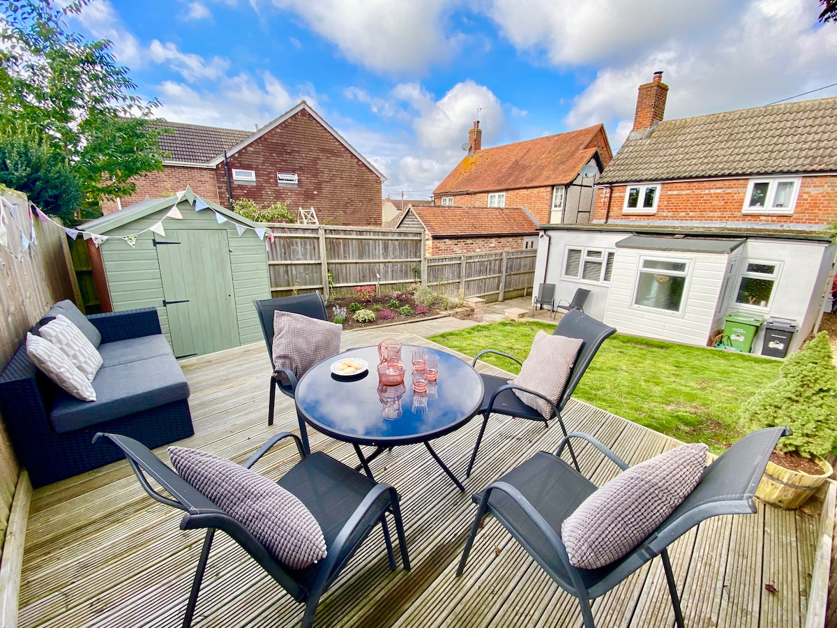 A spacious outdoor deck features a round table surrounded by six chairs, inviting relaxation. A selection of glasses rests atop the table. A shed and well-maintained garden area are visible in the background, with charming houses completing the scenic view.