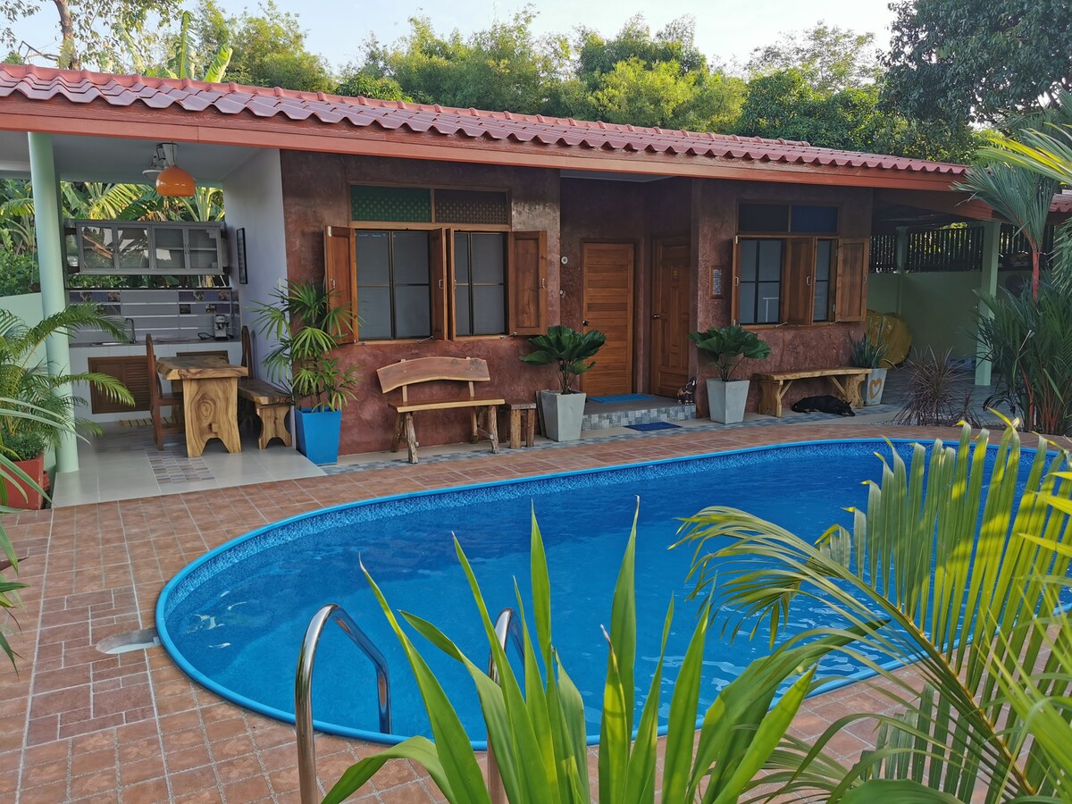 A private guesthouse is surrounded by lush greenery, featuring a shimmering pool directly in front. The structure offers a cozy outdoor seating area with wooden benches, and large windows allow natural light to fill the inviting interior.